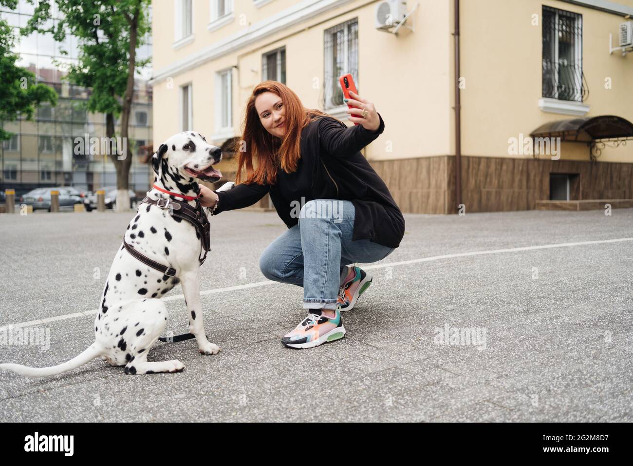 Giovane donna rossastra per strada con il suo cane dalmata scattando una foto con il cellulare. Stile di vita all'aperto con animali domestici. Amore, cura e amicizia Foto Stock