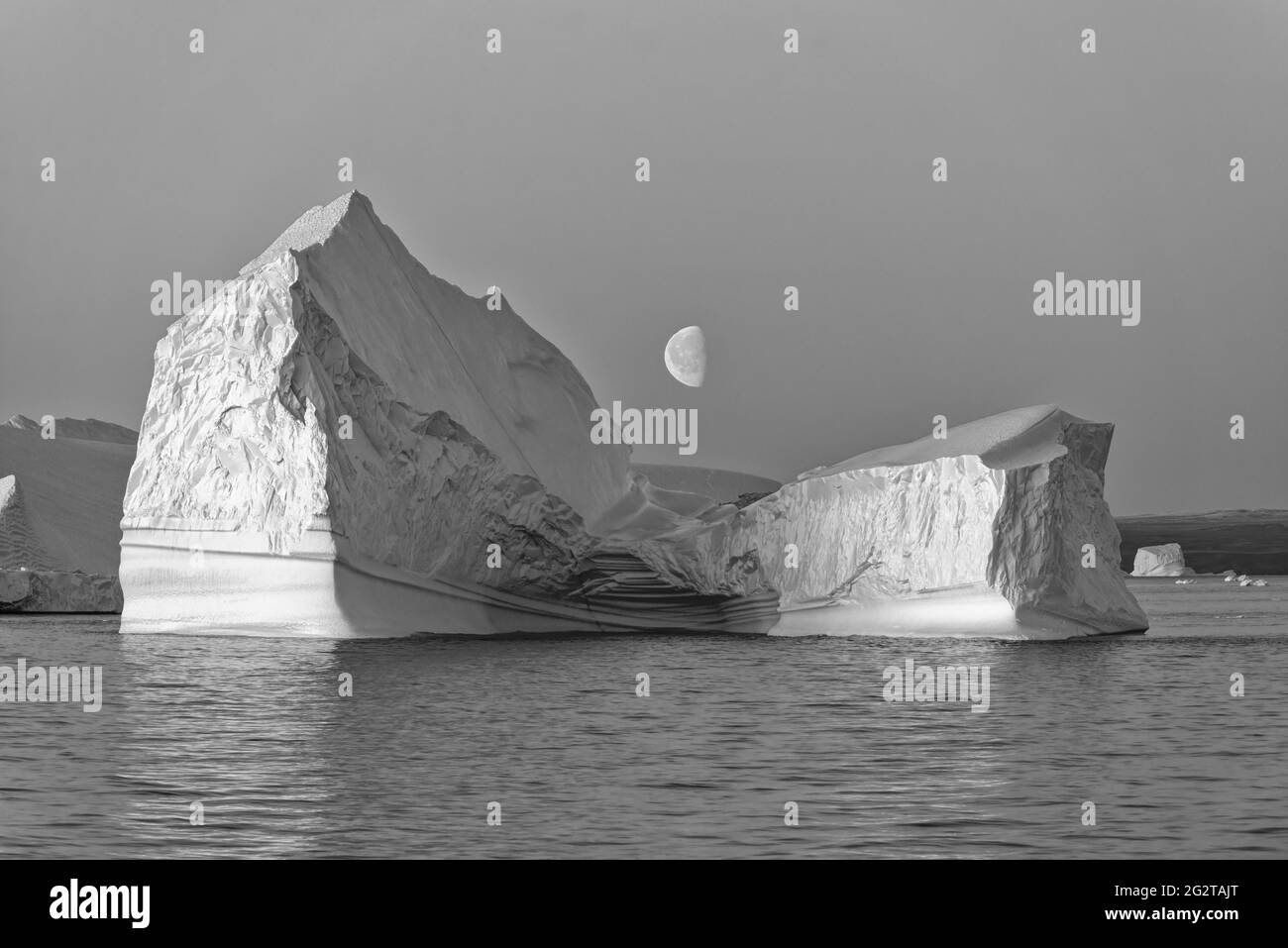 Foto in bianco e nero di un enorme iceberg galleggiante al crepuscolo con la luna nel mezzo, Scoresby Sund, Kangertittivaq, Groenlandia, Danimarca Foto Stock