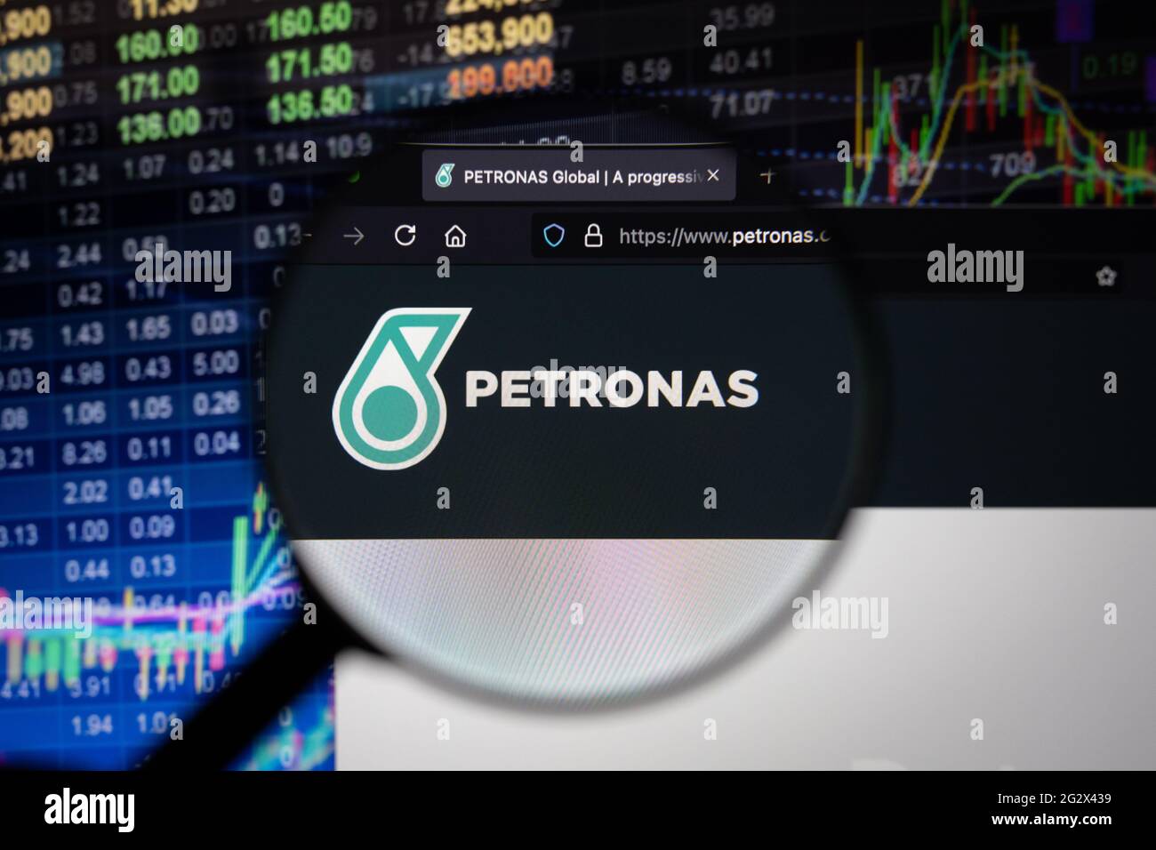 Logo aziendale Petronas su un sito Web con sviluppi sfocati del mercato azionario in background, visto su uno schermo del computer attraverso una lente di ingrandimento Foto Stock