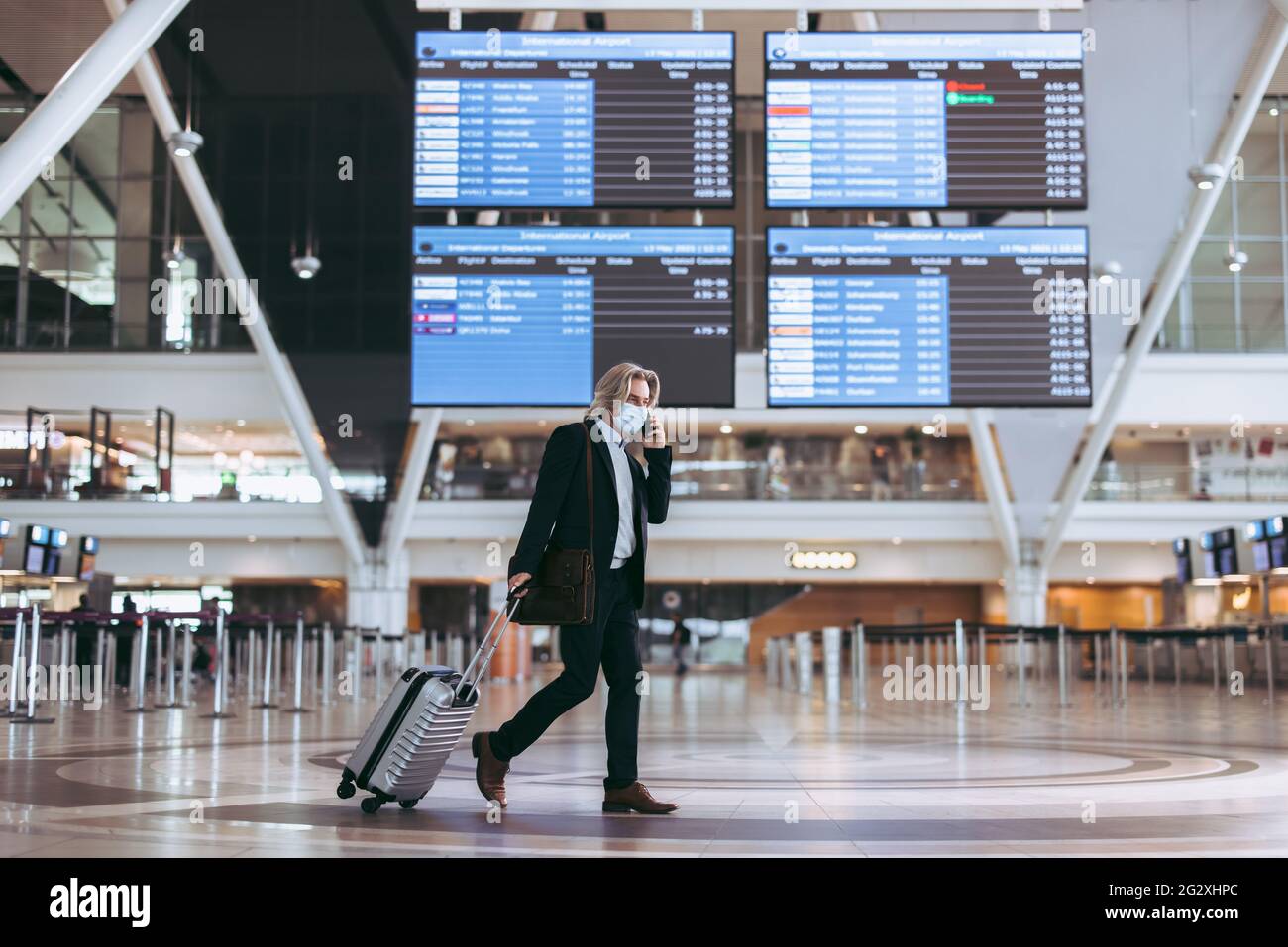 Uomo d'affari maturo che indossa una maschera che parla al telefono mentre si trova al terminal dell'aeroporto. Uomo in viaggio d'affari dopo blocco pandemico. Foto Stock