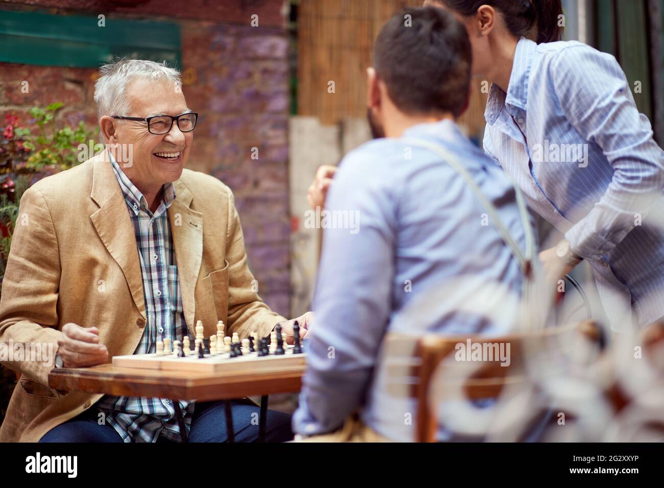 il vecchio maschio caucasico sorridente, felice di vincere nel gioco degli scacchi. seduto in un caffè all'aperto con il suo avversario più giovane che sta parlando con una donna. Foto Stock