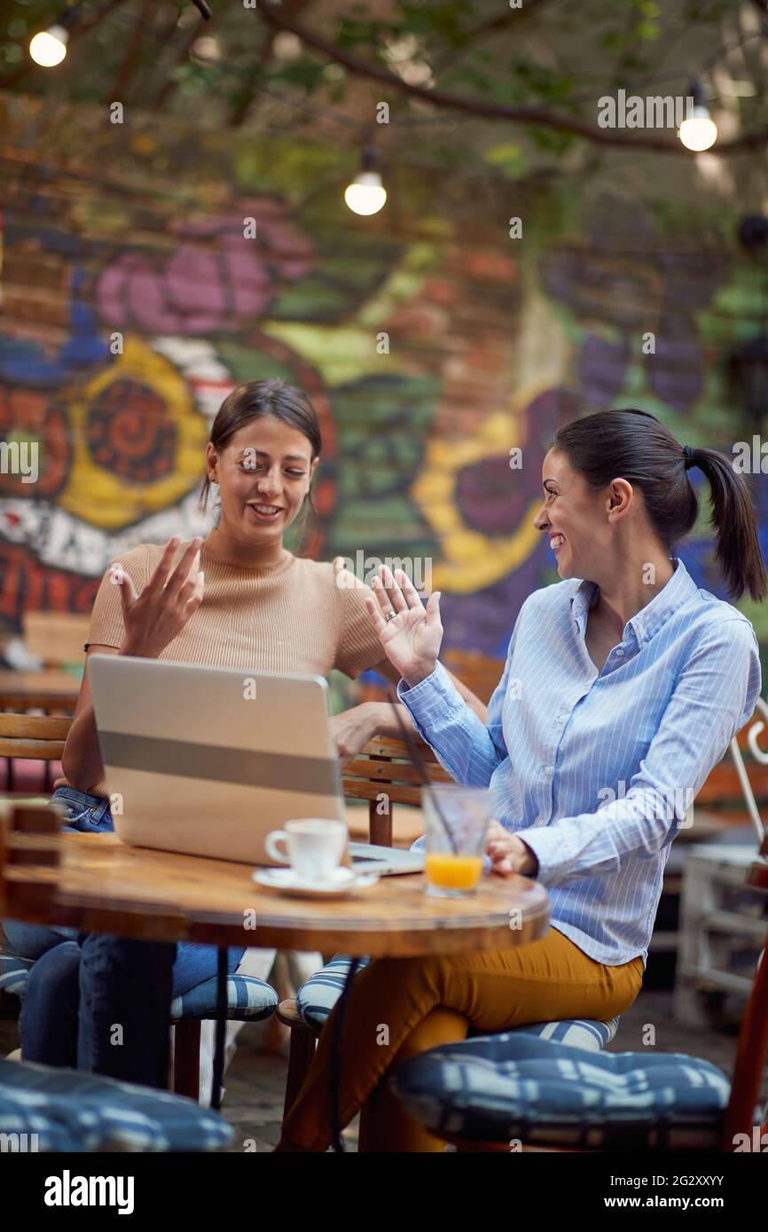 due giovani donne caucasiche adulte sedute in un caffè all'aperto, sorridenti, con palme nell'aria che si preparano per cinque Foto Stock