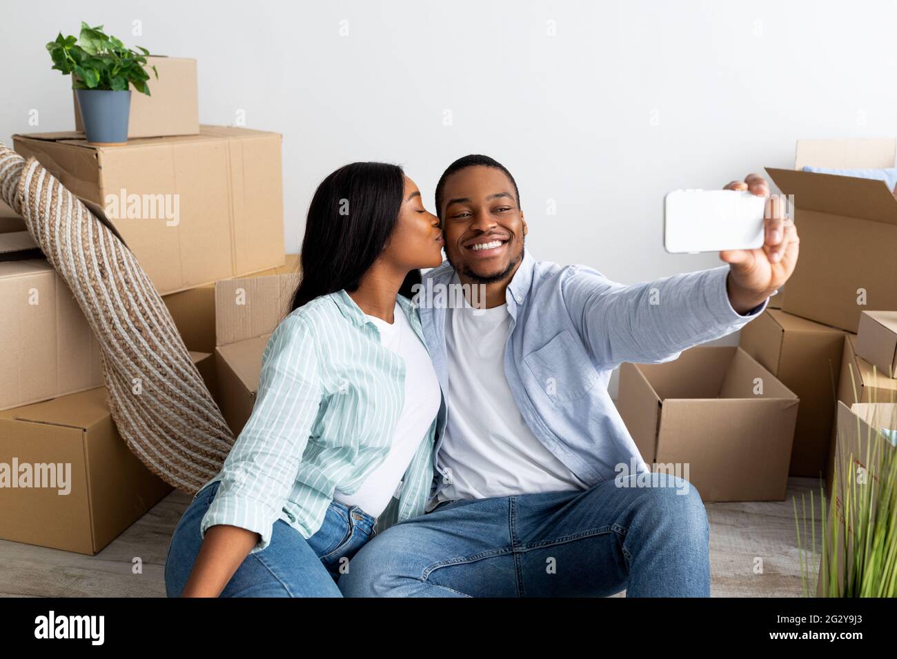 Amorevoli sposi neri che si spostano a casa propria, prendendo selfie tra scatole di cartone, moglie che bacia marito in guancia Foto Stock