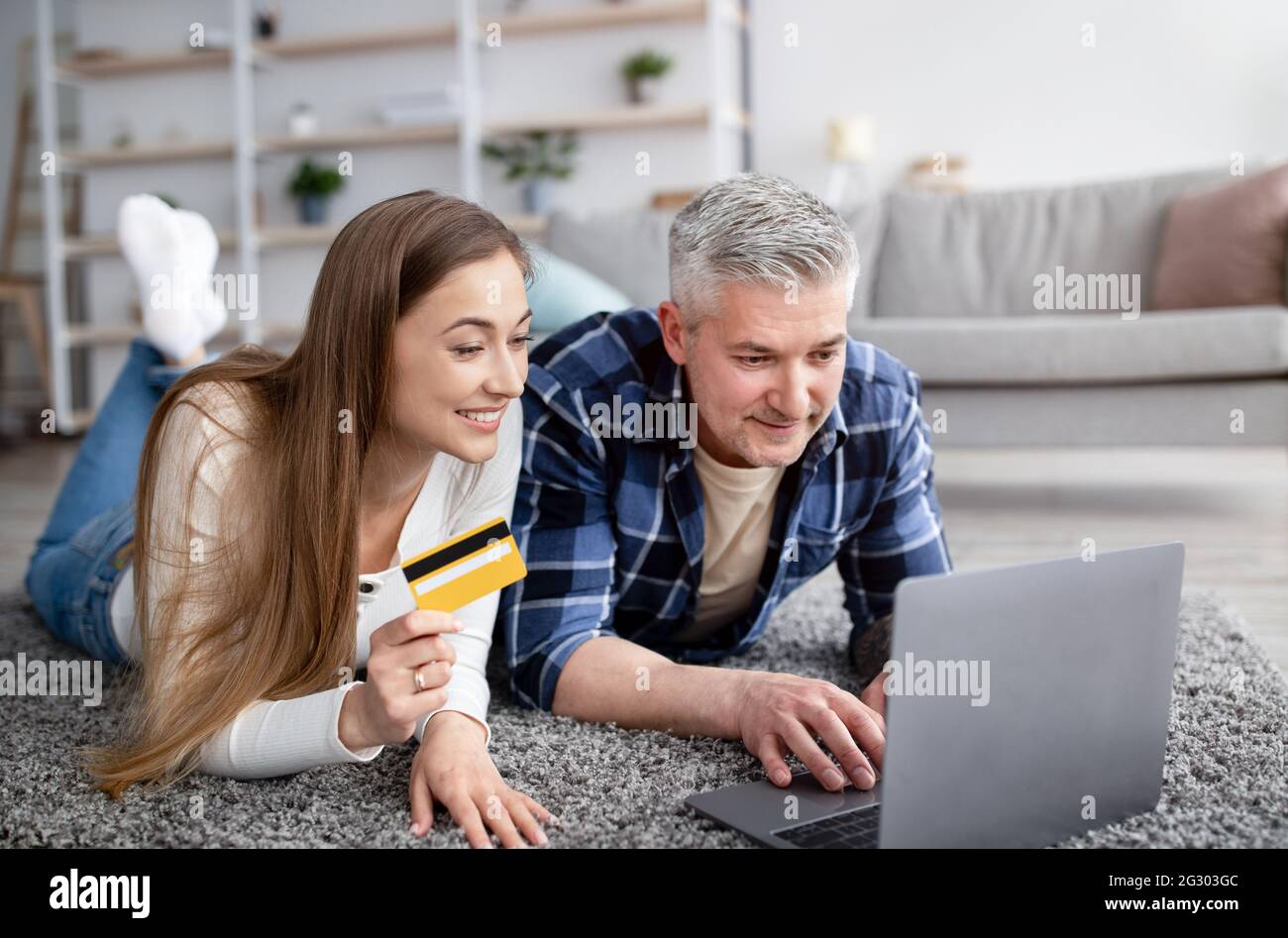 Allegro coniugi maturi che ordinano le merci on-line con PC portatile e carta di credito, sdraiati sul pavimento a casa Foto Stock