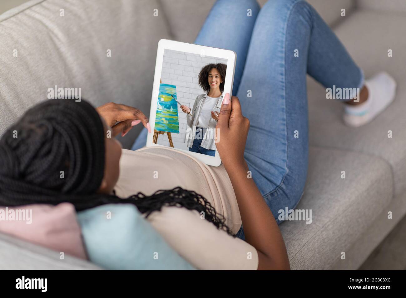 Donna nera irriconoscibile sdraiata sul divano a casa e che guarda la lezione di pittura online sul tablet Foto Stock