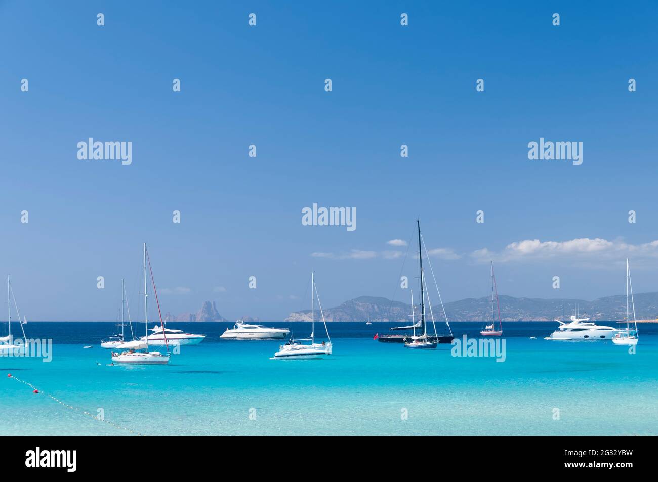 Varie barche a vela ormeggiate nel mare turchese vicino ad una spiaggia sull'isola di Formentera. Sullo sfondo il cielo blu e la costa di Ibiza Foto Stock