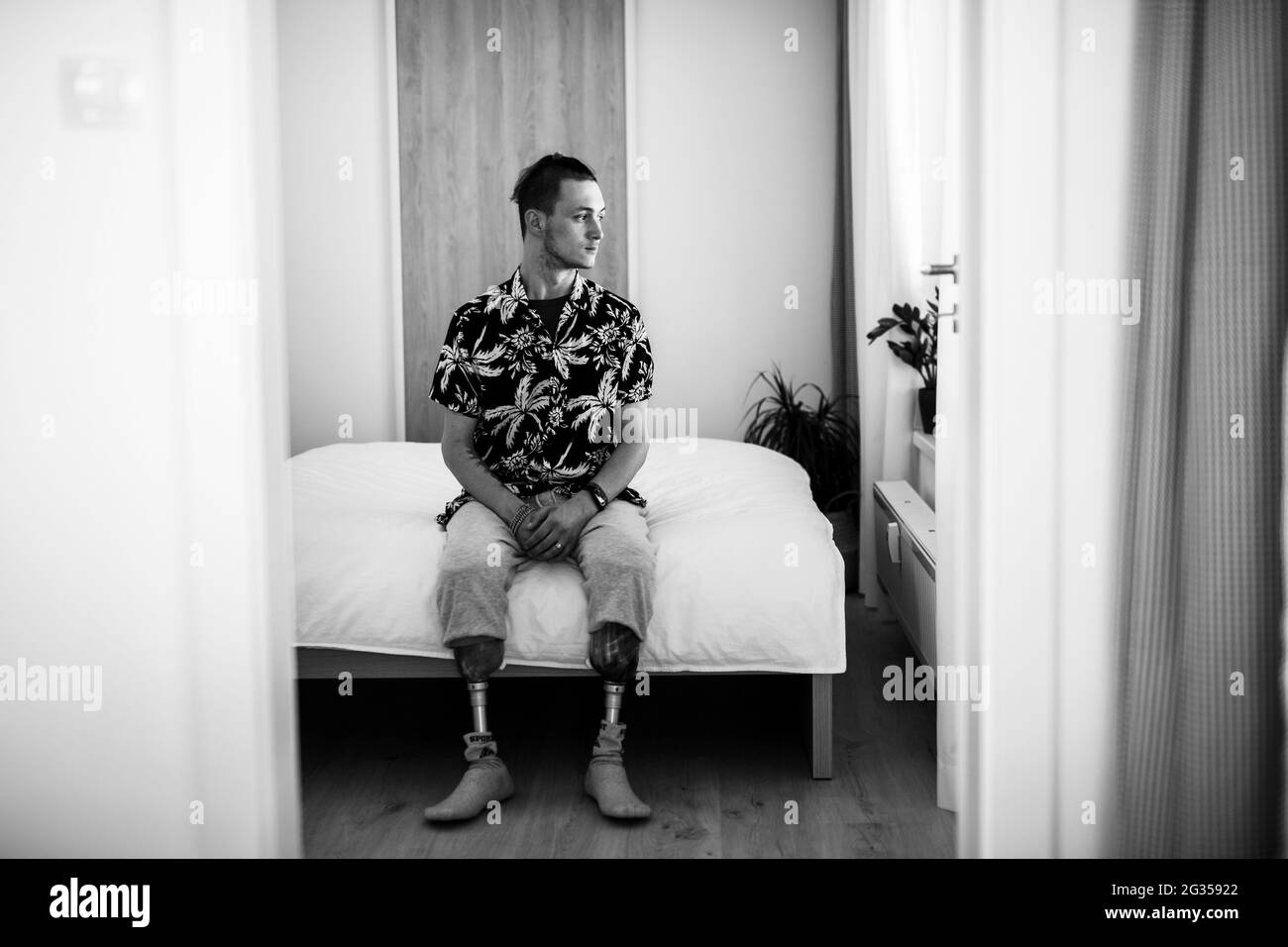 Ritratto in bianco e nero di un giovane disabile seduto a letto in casa, concetto protesico delle gambe. Foto Stock