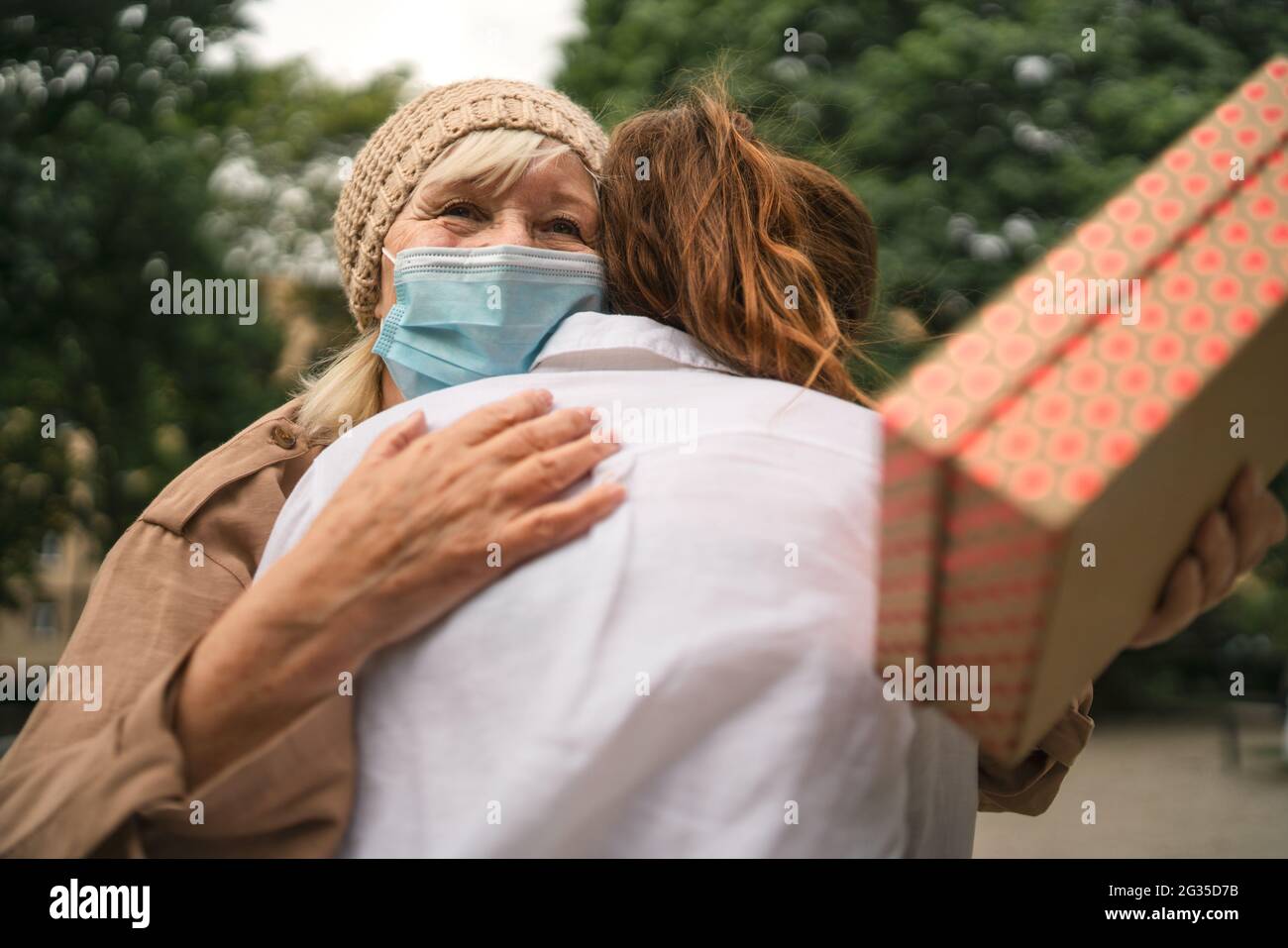 Giovane donna che dà presente e abbracciando nonna all'aperto in città, la vita dopo la vaccinazione covid-19. Foto Stock