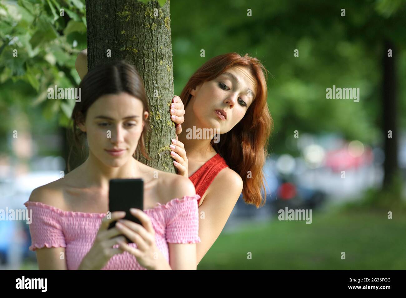 Donna distratta che usa lo smartphone che viene spiata dal suo amico curioso in un parco Foto Stock