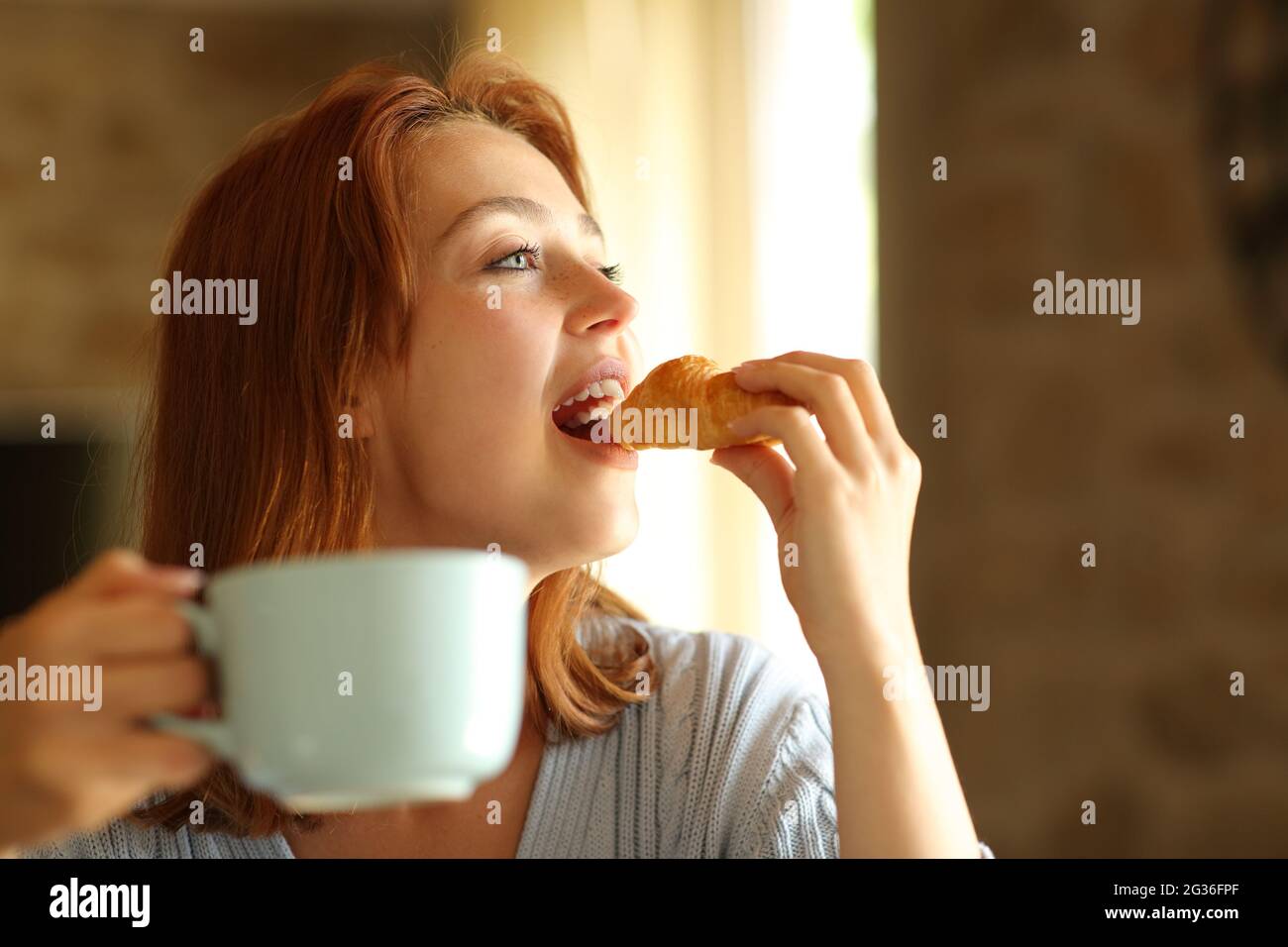Donna felice che mangia e beve per la prima colazione nel soggiorno di casa Foto Stock