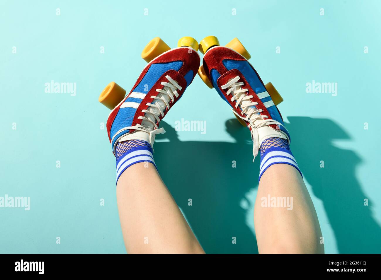 POV in prima persona delle gambe di una donna che indossa colorati pattini a rotelle rosso e blu con ruote gialle e calzini su uno sfondo blu studio Foto Stock