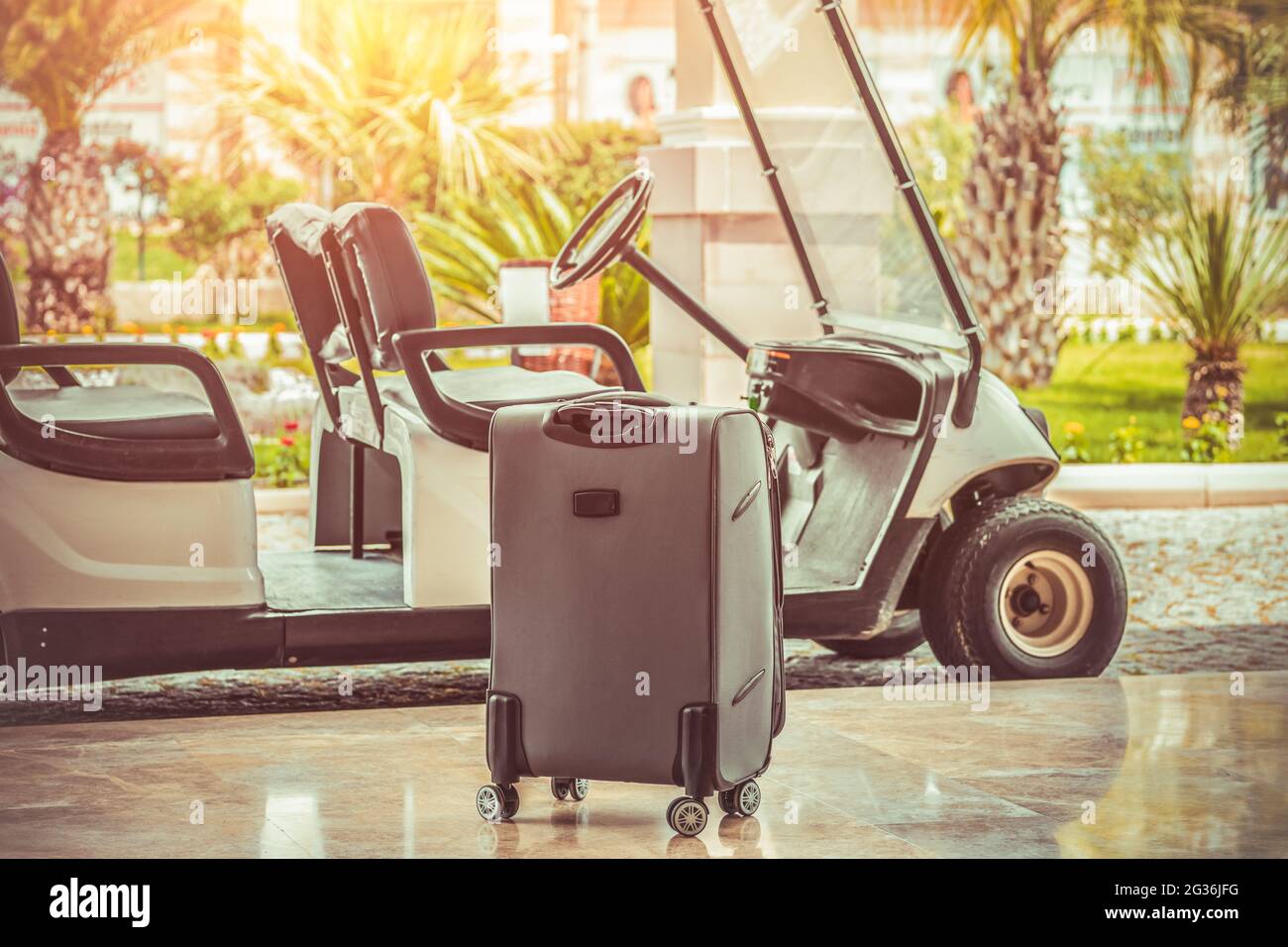 Una valigia in piedi vicino all'autobus navetta per passeggeri dell'hotel elettrico di fronte a palme sfocate. Concetto di vacanza estiva con servizio di auto da golf e bagagli. Foto Stock