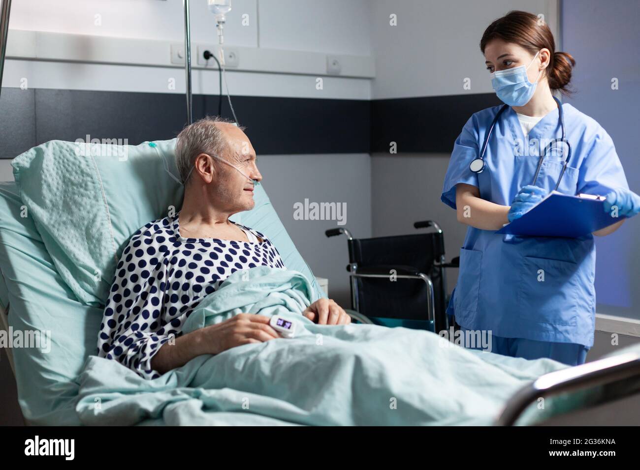 Infermiera medica in scrub con maschera chirurgica che prende notest sulla scacchiera durante la consultazione di malato uomo anziano malato. Paziente con respirazione fleboclisi ev con l'aiuto della maschera di ossigeno. Foto Stock