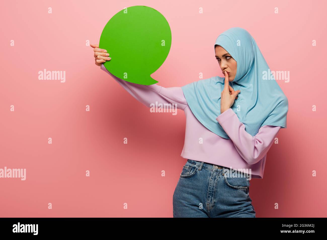 donna musulmana premurosa che tocca le labbra mentre si guarda la bolla di pensiero verde su sfondo rosa Foto Stock