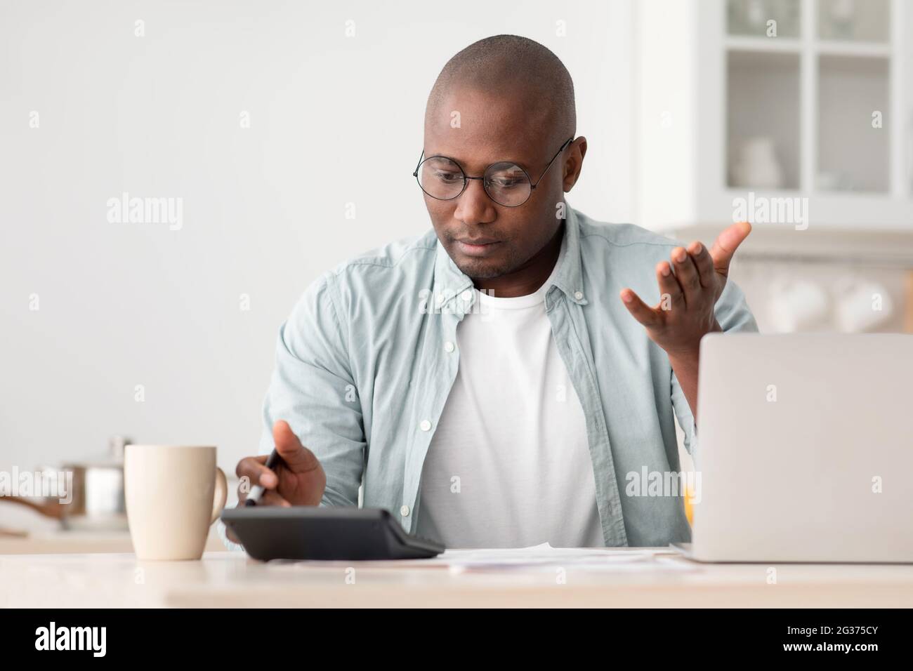 Concetto di risparmio e finanze. Uomo nero scioccato maturo calcolando le fatture, utilizzando la calcolatrice, seduto in cucina Foto Stock