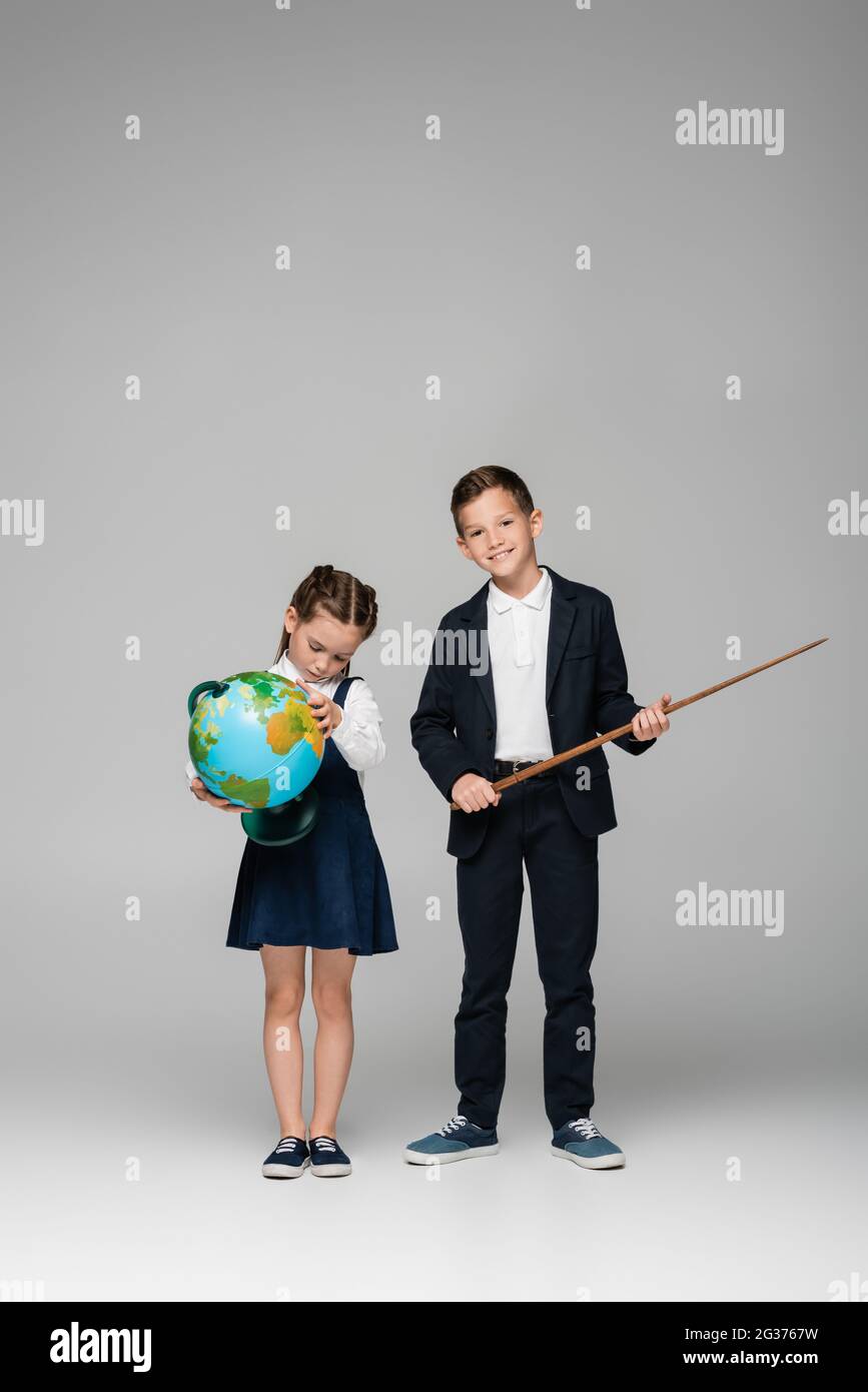 cheerful schoolboy che tiene il bastone di puntamento vicino alla ragazza triste in vestito con il globo su grigio Foto Stock