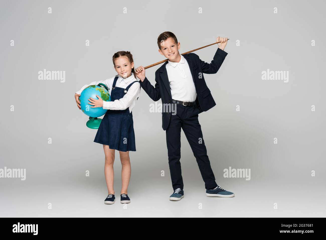 felice scolaro che tiene il bastone di puntamento vicino alla ragazza sorridente in abito con il globo su grigio Foto Stock