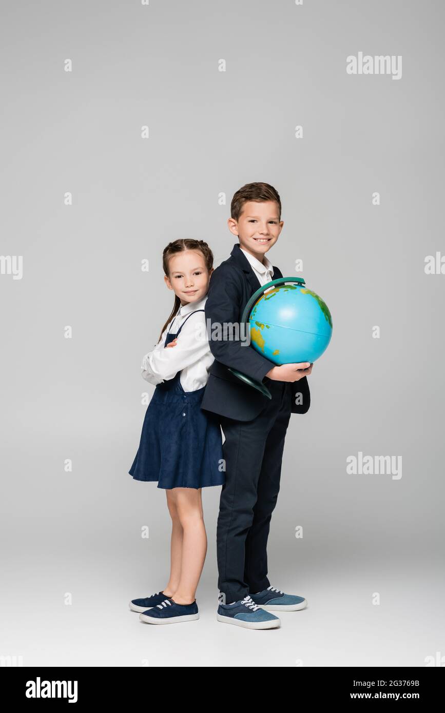 felice scolaro tenendo il globo mentre si sta con la ragazza in abito su grigio Foto Stock