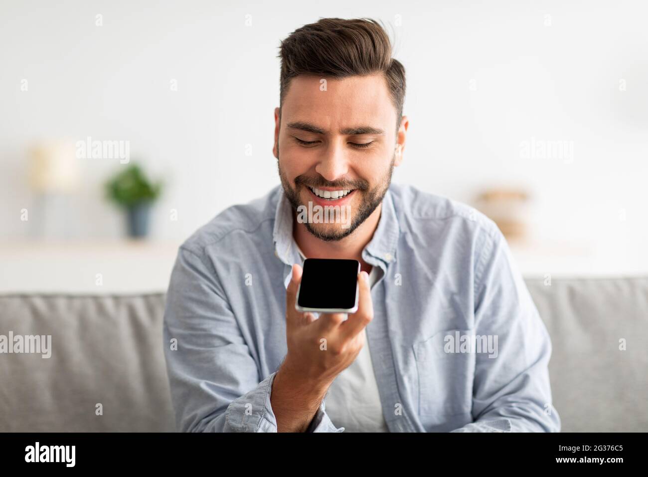 Uomo felice bello che registra un messaggio vocale sullo smartphone o utilizza un assistente virtuale mentre si riposa sul divano a casa Foto Stock