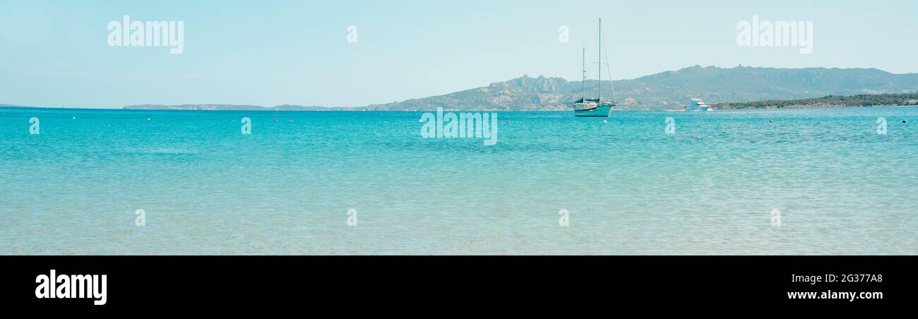 Una barca a vela e uno yacht stanno navigando sulle acque calme del Mediterraneo in Sardegna, Italia, visto dalla Spiaggia di Vena Longa, in un panoramico per Foto Stock