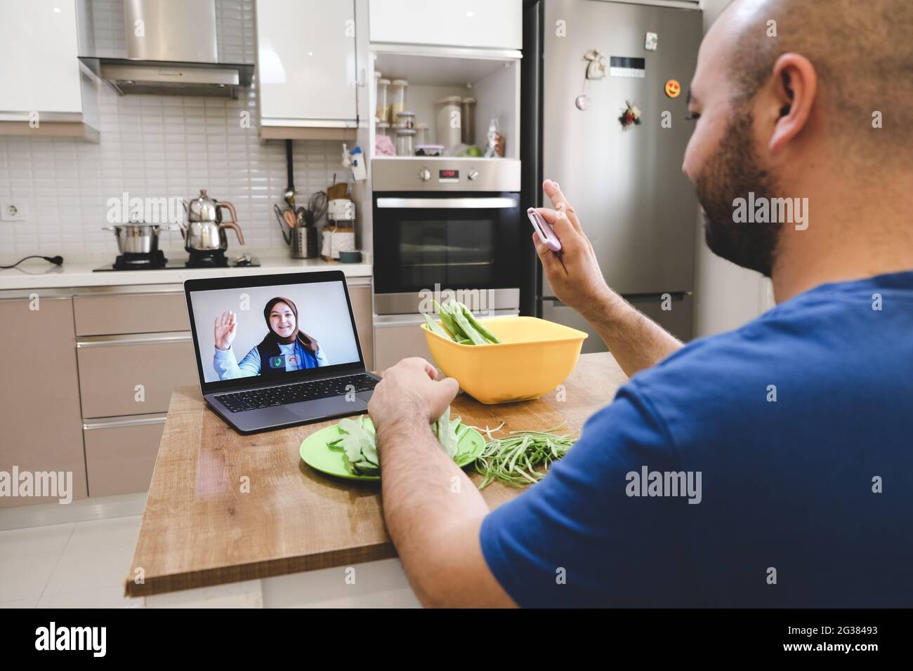 Persone che salutano in videoconferenza mentre preparano un piatto di verdure in cucina. Donna e uomo con videochiamata online. Foto Stock