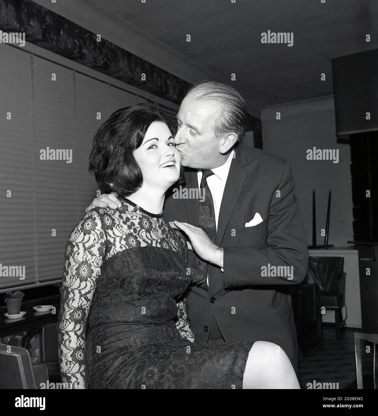 1965, storica, una graziosa giovane donna dai capelli scuri, il vincitore del concorso di bellezza 'Maid of Loch' che ottiene un bacio di congratulazioni o un pick sulla guancia da un gentleman ben vestito, casa Brucefield, Scozia, Regno Unito. Foto Stock