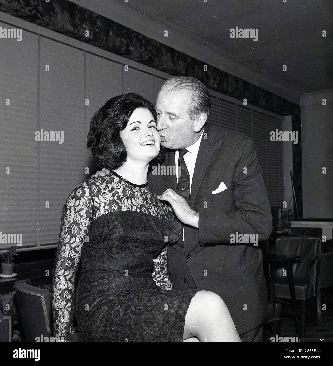 1965, storica, una graziosa giovane donna dai capelli scuri, il vincitore del concorso di bellezza 'Maid of Loch' che ottiene un bacio di congratulazioni o un pick sulla guancia da un gentleman ben vestito, casa Brucefield, Scozia, Regno Unito. Foto Stock