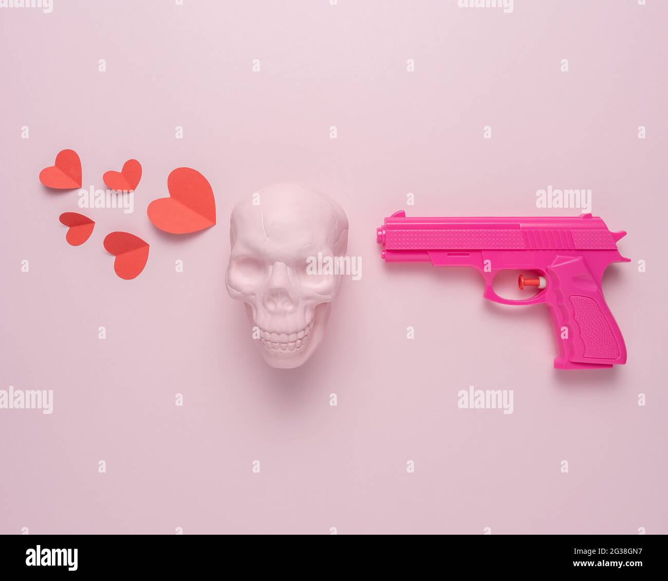 Pistola rosa su sfondo rosa pastello con cranio rosa e cuori di carta rossa. Concetto di amore minimo. Foto Stock