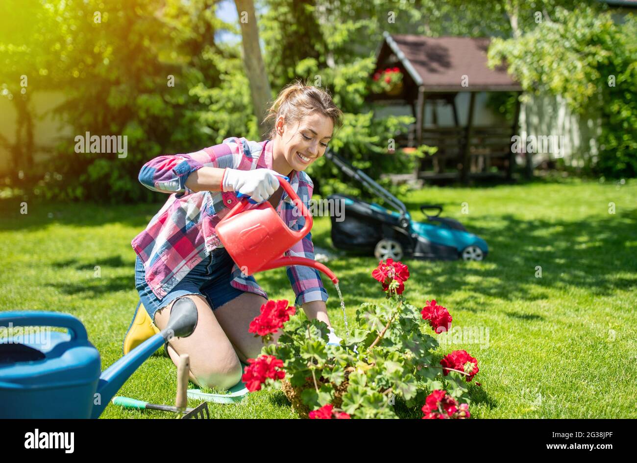 Felice giovane donna giardinaggio in giornata di sole di fronte al tosaerba. Giardiniere inginocchiato su erba che annaffia geranio in fiore. Foto Stock