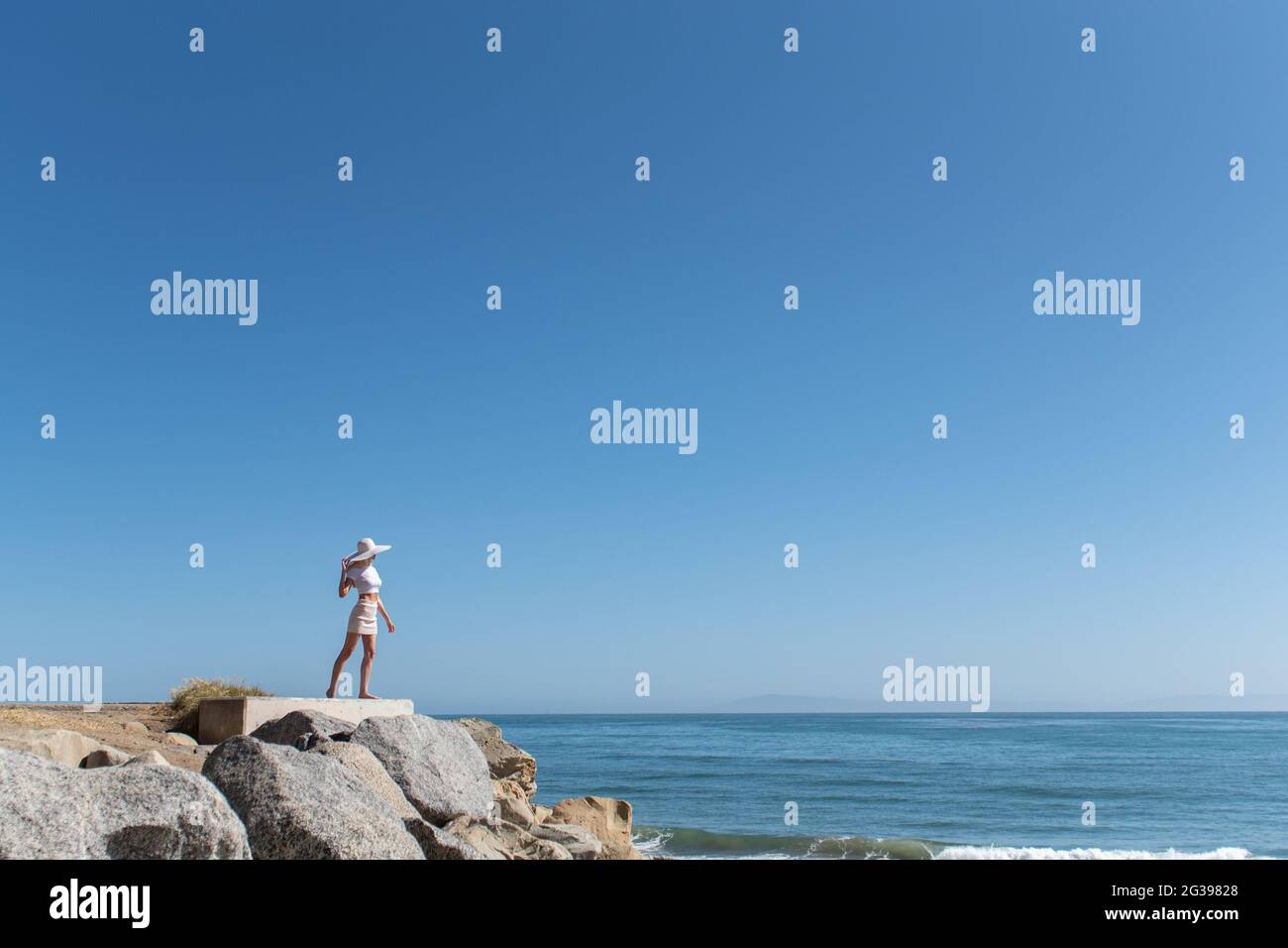 Bella modella femmina dei capelli biondi aggrappato al cappello mentre guardando fuori nel grande Oceano Pacifico dalla costa rocciosa. Foto Stock