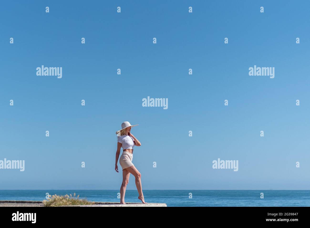 Bella modella femmina dei capelli biondi aggrappato al cappello mentre guardando fuori nel grande Oceano Pacifico con orizzonte in linea giù alle sue caviglie. Foto Stock