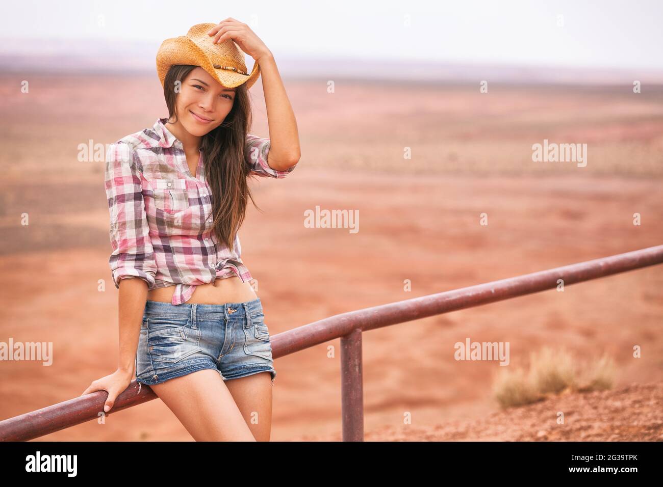 Donna Cowgirl sorridente a macchina fotografica che tiene il cappello come gesto di saluto felice sul paesaggio agricolo di campagna che indossa cowboy occidentale camicia e jeans. Giovane Foto Stock