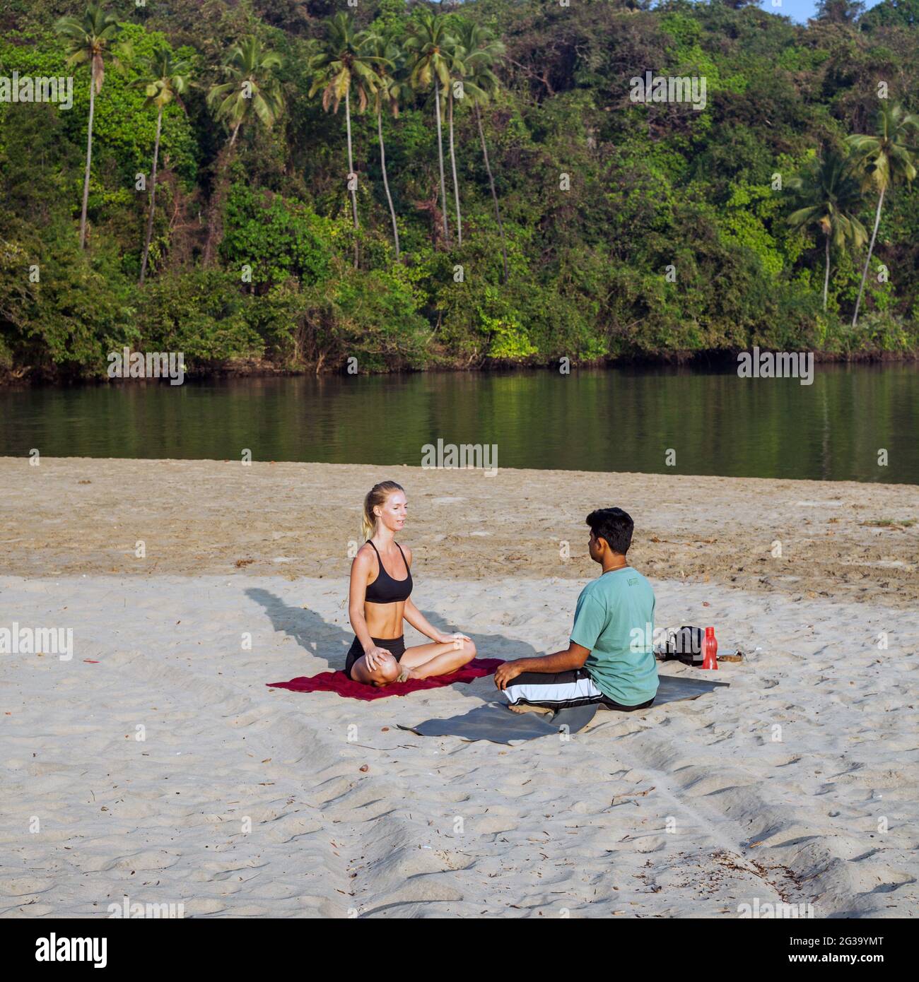 Bella bionda occidentale seduto in posizione lotus pratica yoga sulla spiaggia dopo l'alba con il suo ragazzo indiano, Agonda, Goa, India Foto Stock