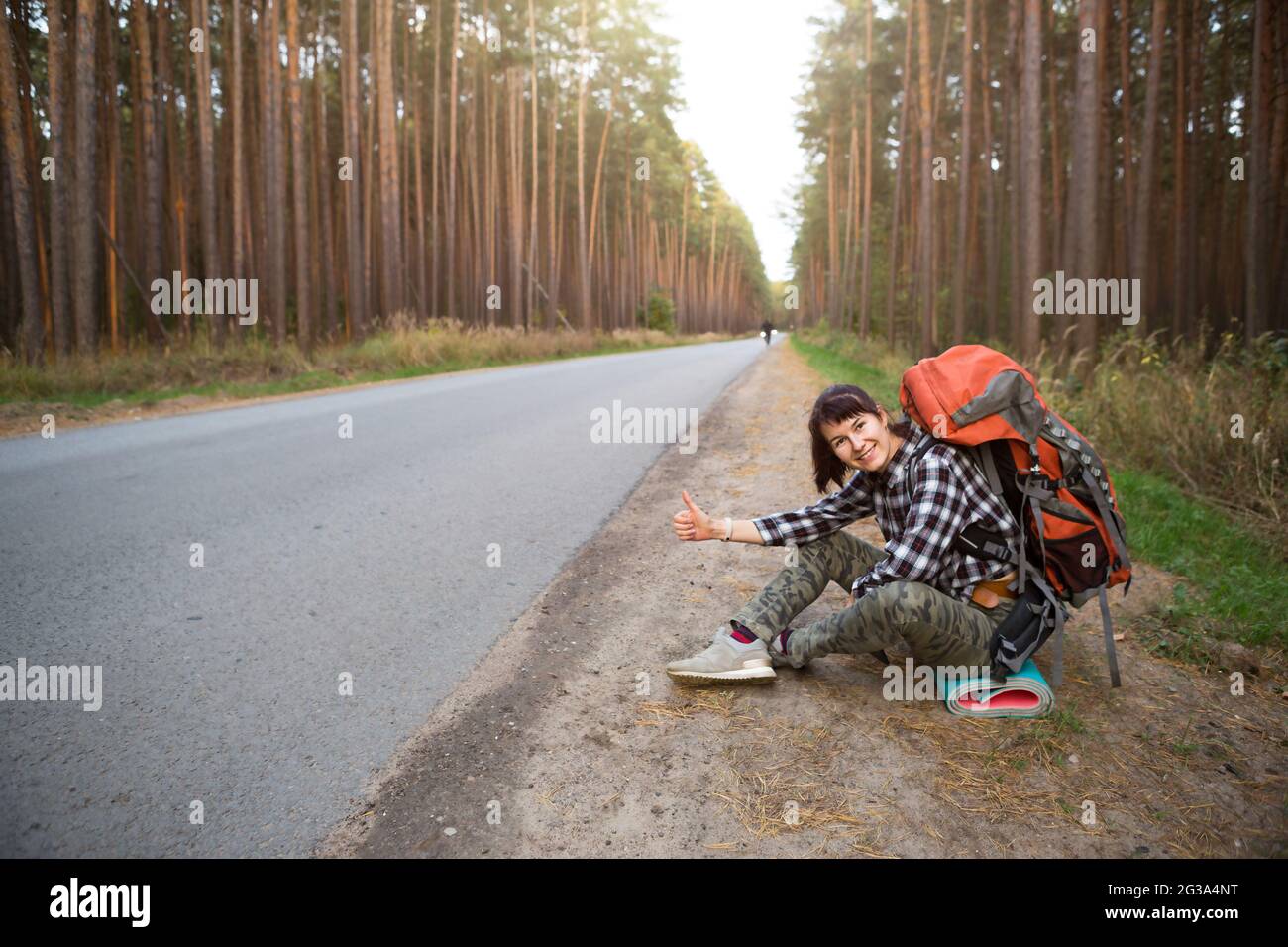 Donna turistica in una camicia controllata con uno zaino arancione grande vicino a un'autostrada nel bosco voti per ottenere un giro. Escursioni, turismo domestico. Backpa Foto Stock