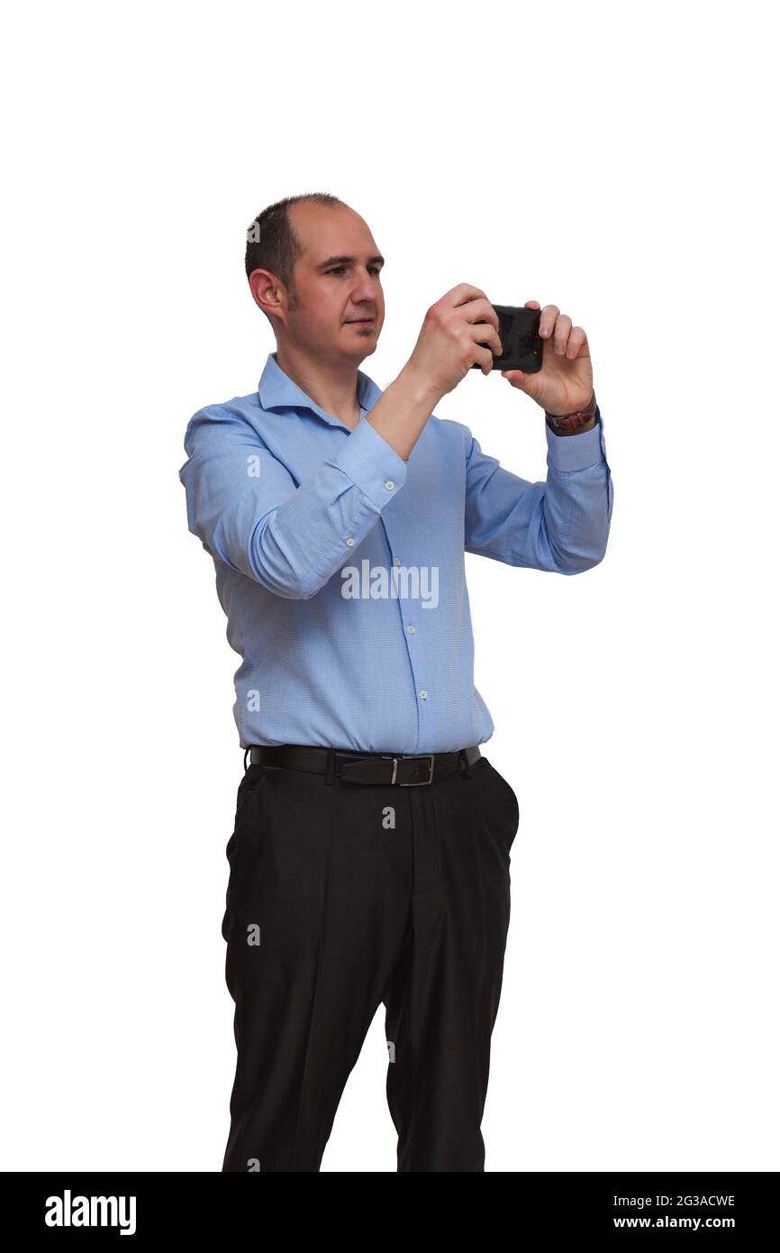 Uomo calvo vestito con camicia blu e pantaloni neri che scatta una foto o registra un video con il suo smartphone isolato su sfondo bianco. Può essere un ufficio WO Foto Stock