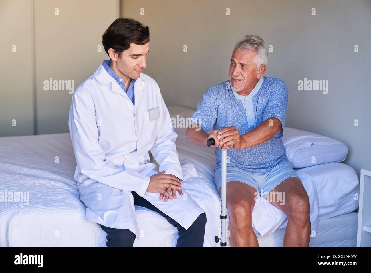 Anziano con aiuto ambulante siede sul letto e parla con un'infermiera geriatrica nella casa di cura Foto Stock