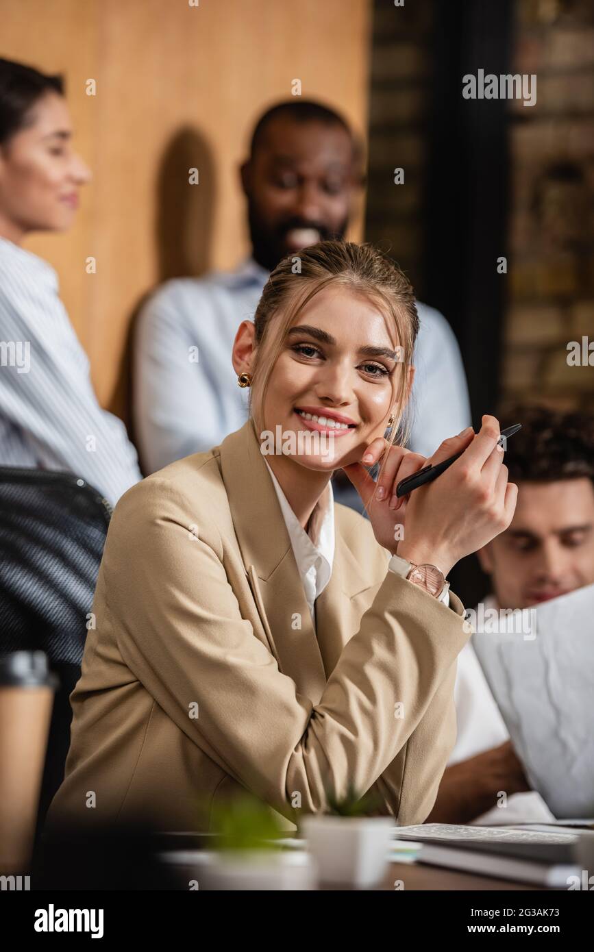donna d'affari gioiosa che guarda la macchina fotografica vicino i soci interracial offuscati di affari Foto Stock
