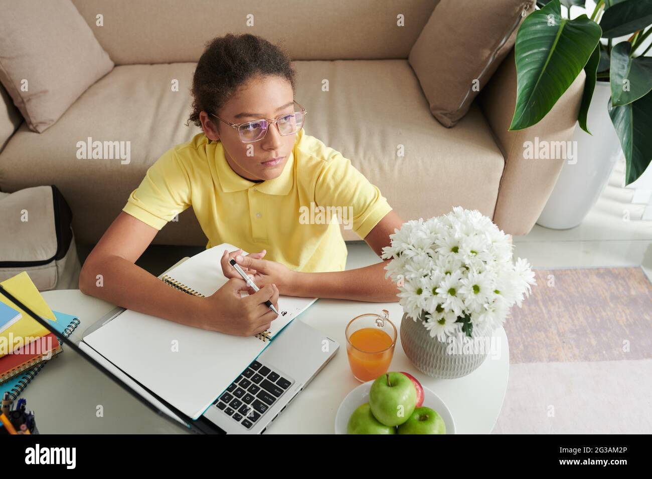 Ragazza adolescente di razza mista di pene in bicchieri seduti a tavolino con fiori in vaso e utilizzando il computer portatile mentre prendendo note su articolo online Foto Stock