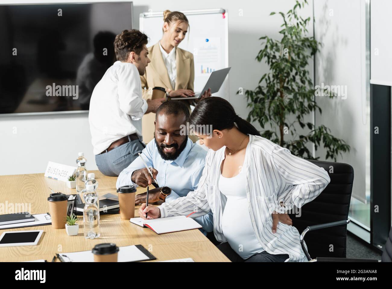 sorridente afroamericano che punta al notebook vicino donna incinta d'affari e colleghi parlanti Foto Stock