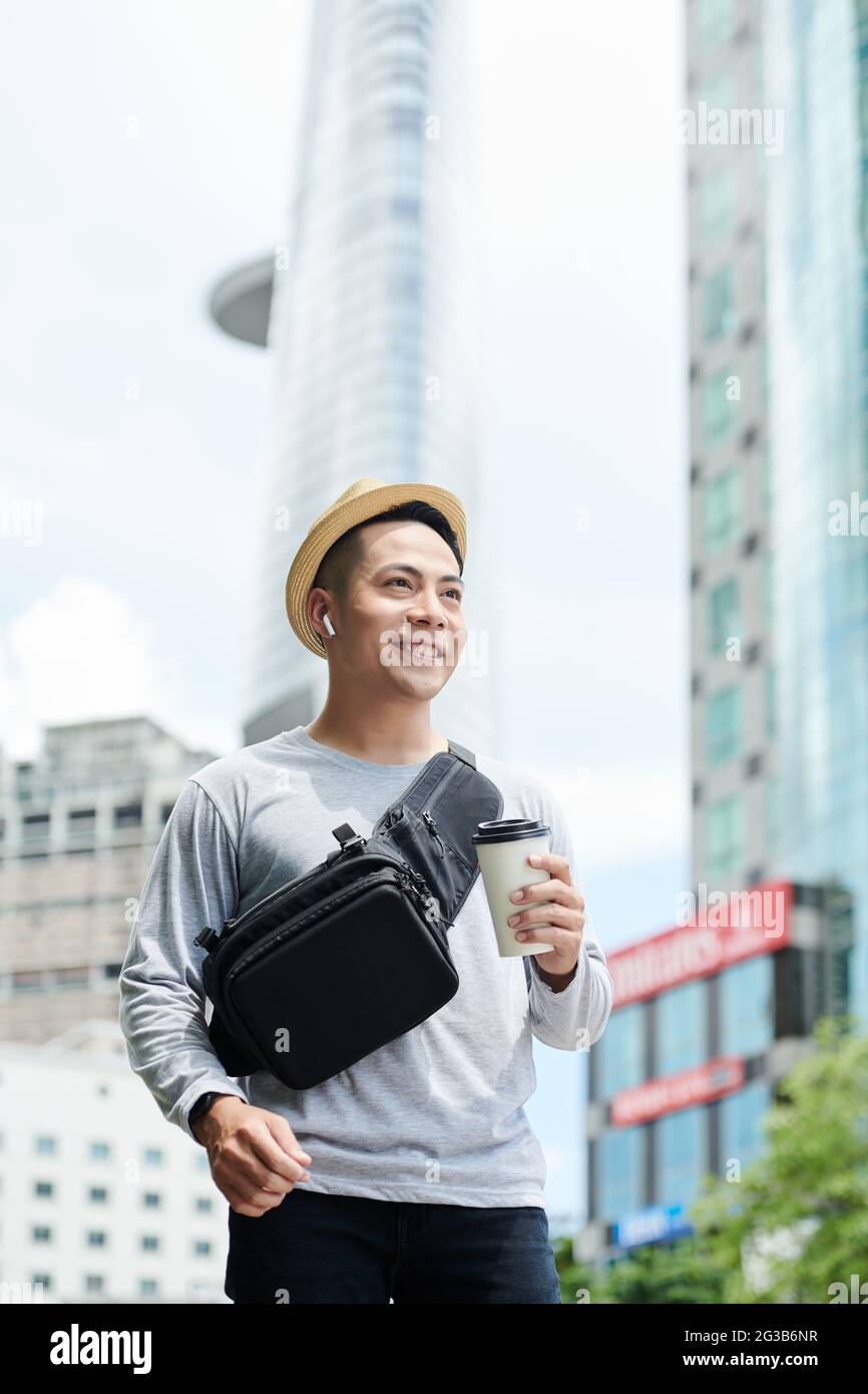 Positivo bell'uomo asiatico in cappello da sole e auricolari wireless che tengono una tazza di caffè e che cammina nella grande città Foto Stock
