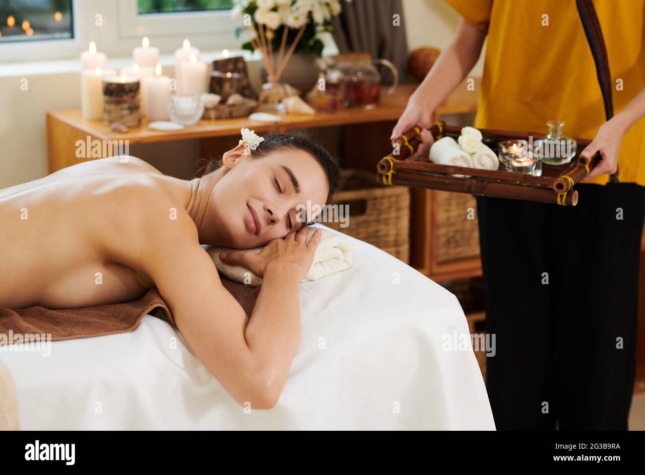 Giovane donna sdraiata sul tavolo da massaggio e rilassante durante la procedura termale con terapista che porta olio aromatico Foto Stock