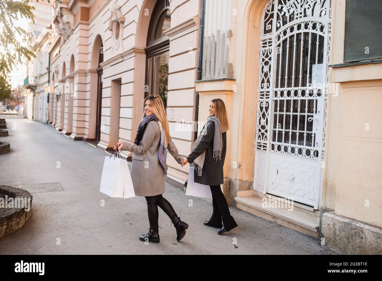 Vista posteriore di due giovani donne bionde che camminano per la città,  tenendo le mani e portando borse per lo shopping. Shopping autunnale nella  città di due ragazze Foto stock - Alamy