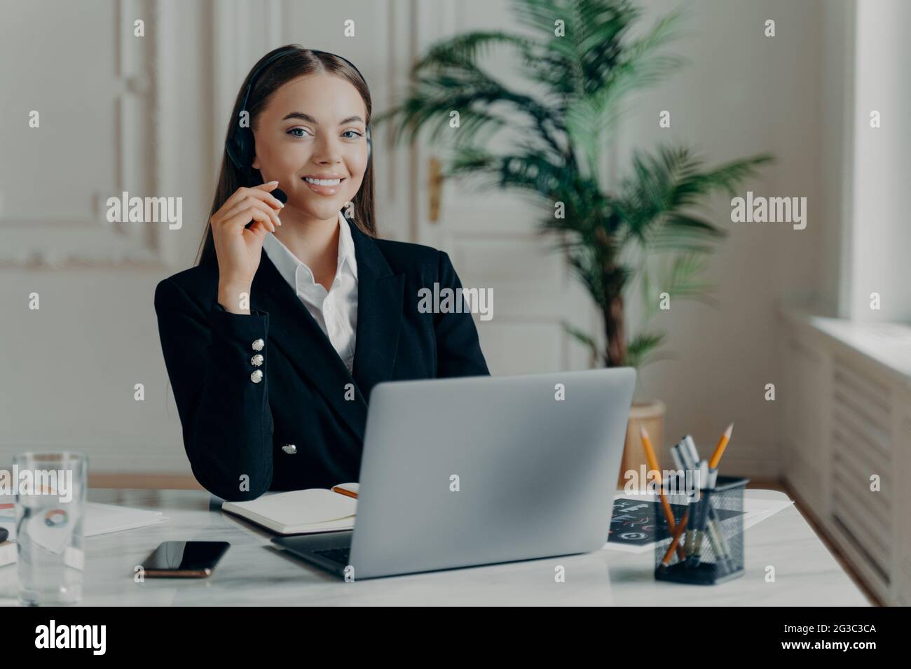 Donna che lavora in ufficio con cuffie sorridenti alla fotocamera durante la riunione online Foto Stock