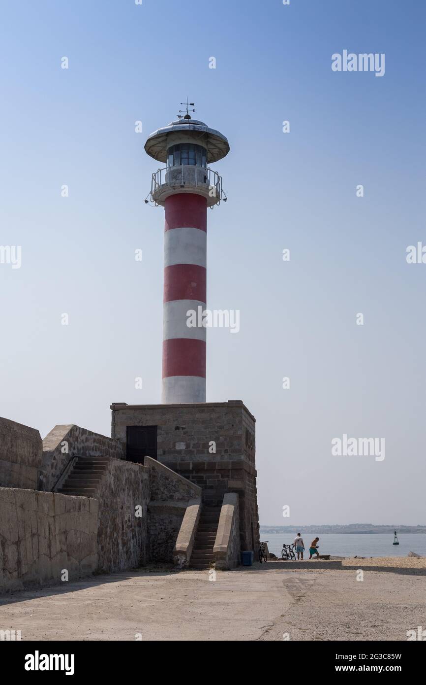 Una vista del porto con un grande vecchio faro rosso bianco, molo contro il cielo blu e il mare Foto Stock