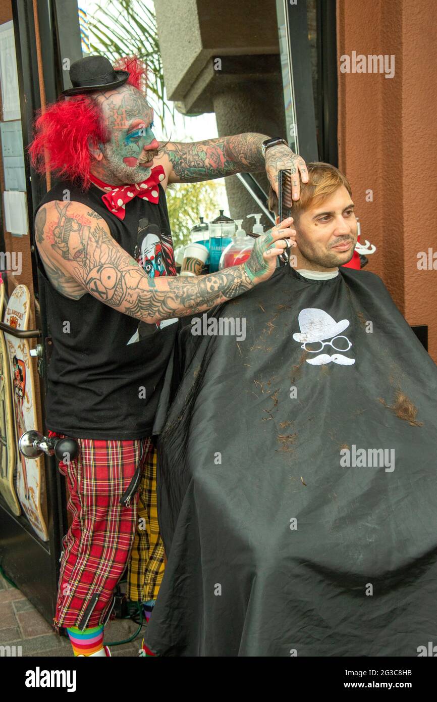 Un barbiere tatuato vestito da pagliaccio rifinisce i capelli di un cliente sul marciapiede a Huntington Beach, CA. Foto Stock