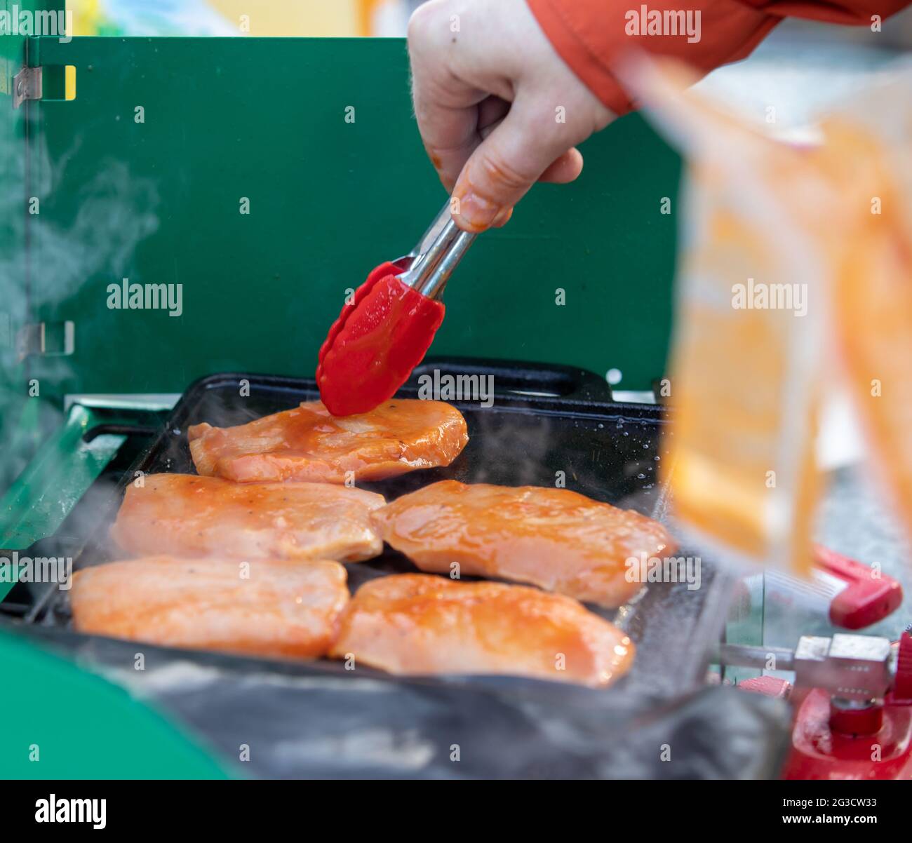 Uomo che tiene piccole pinze da cucina mentre grigliano il pollo marinato su una stufa da campeggio con piastra di ferro Foto Stock