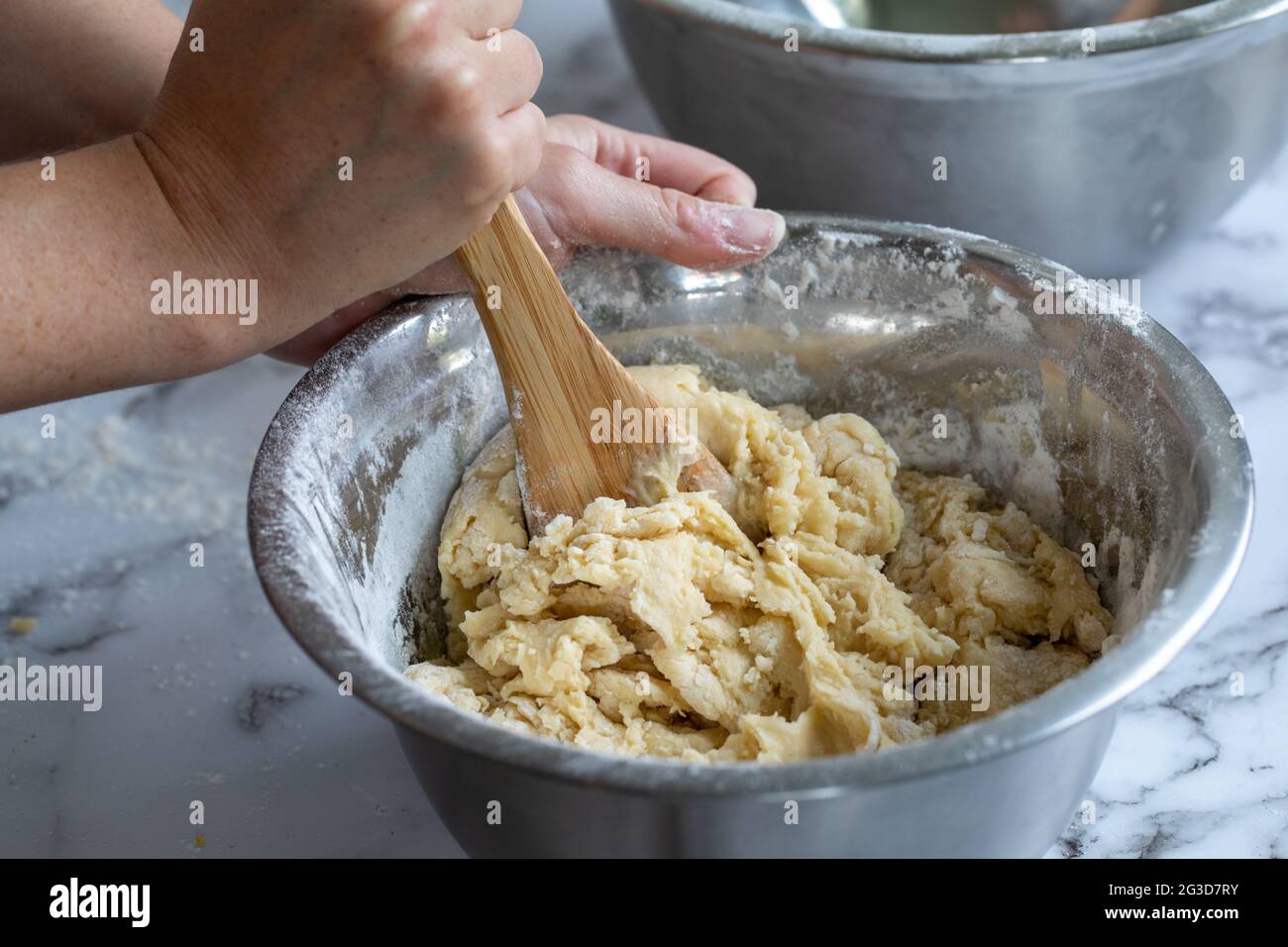 primo piano delle mani di una donna che tiene un cucchiaio di legno mescolando gli ingredienti in una ciotola rotonda di metallo su una superficie di marmo bianco Foto Stock