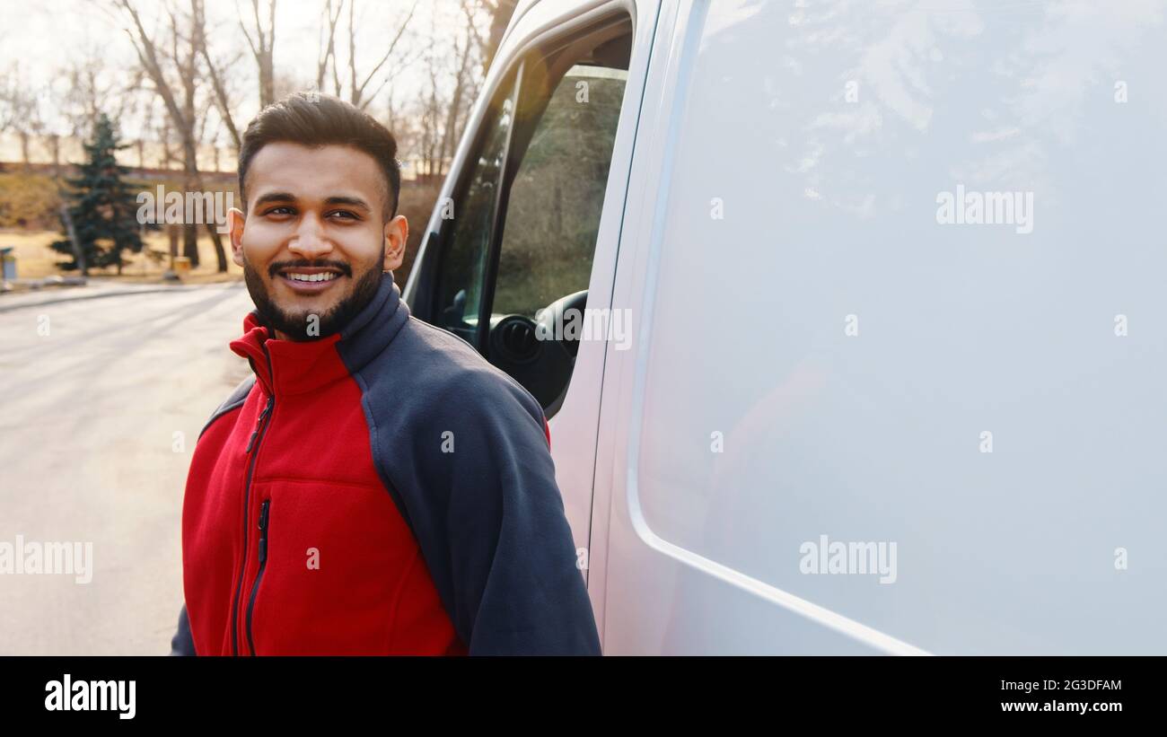Giovane indiano in piedi sulla strada con un furgone bianco dietro di lui. Ragazzo di consegna che indossa uniforme rossa sorridente. Alberi sullo sfondo. Scatto diurno. Foto Stock