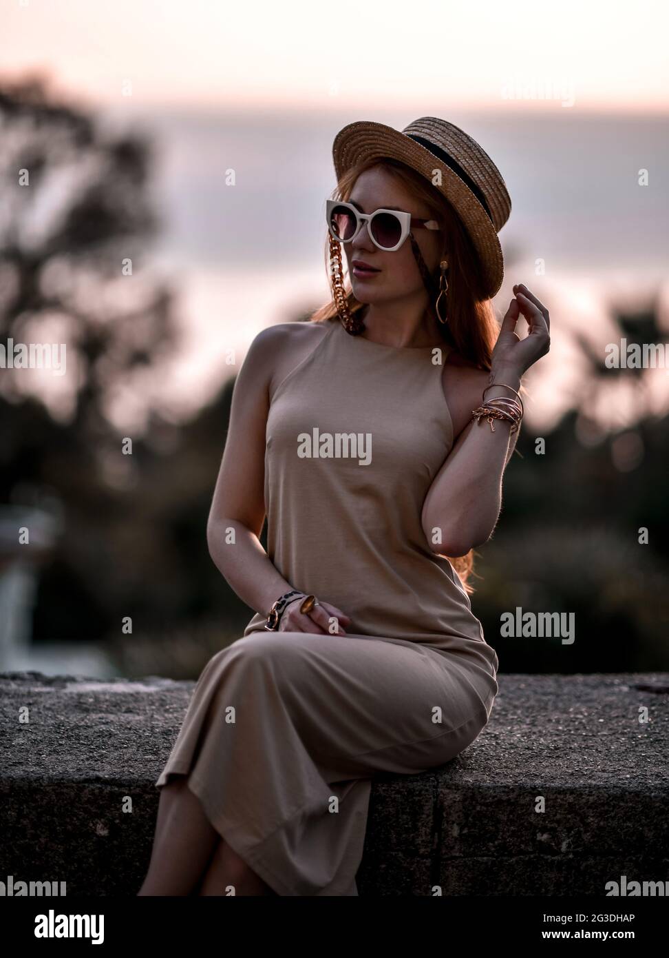 Splendida donna ricca rossa al tramonto estivo seduto su parapetto di pietra in occhiali da sole, cappello di paglia e vestito Foto Stock