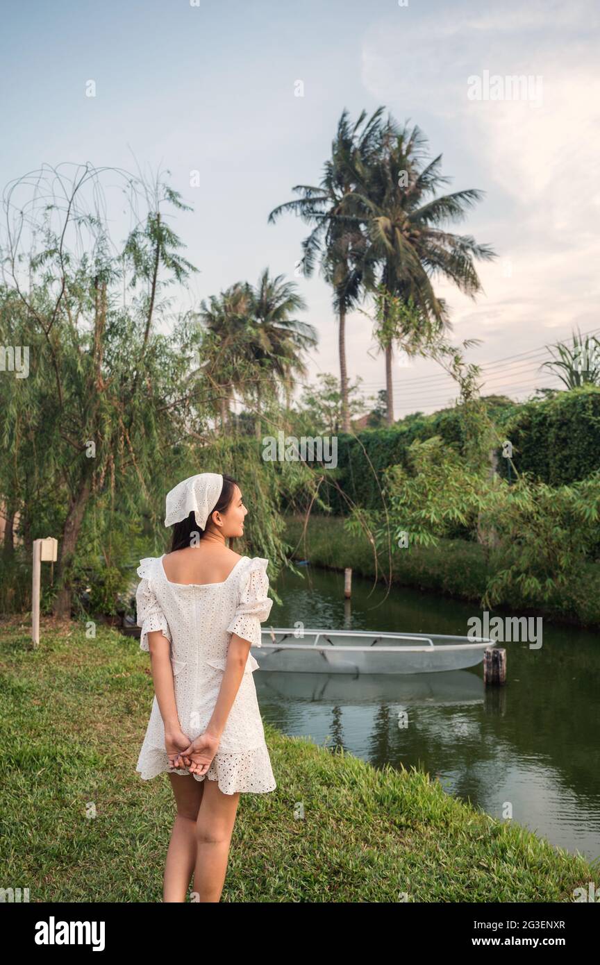 Allegra bella giovane donna asiatica in abito bianco in piedi vicino al canale nel giardino Foto Stock